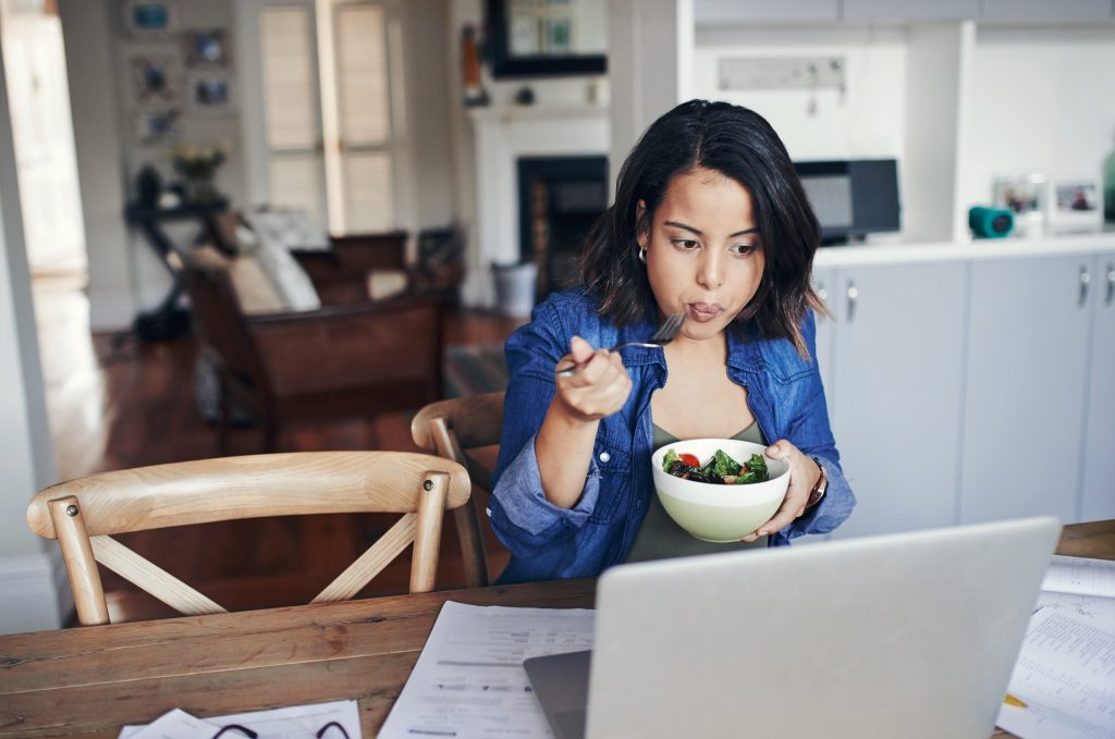 Shot of a young woman using a laptop and having a salad while working from home