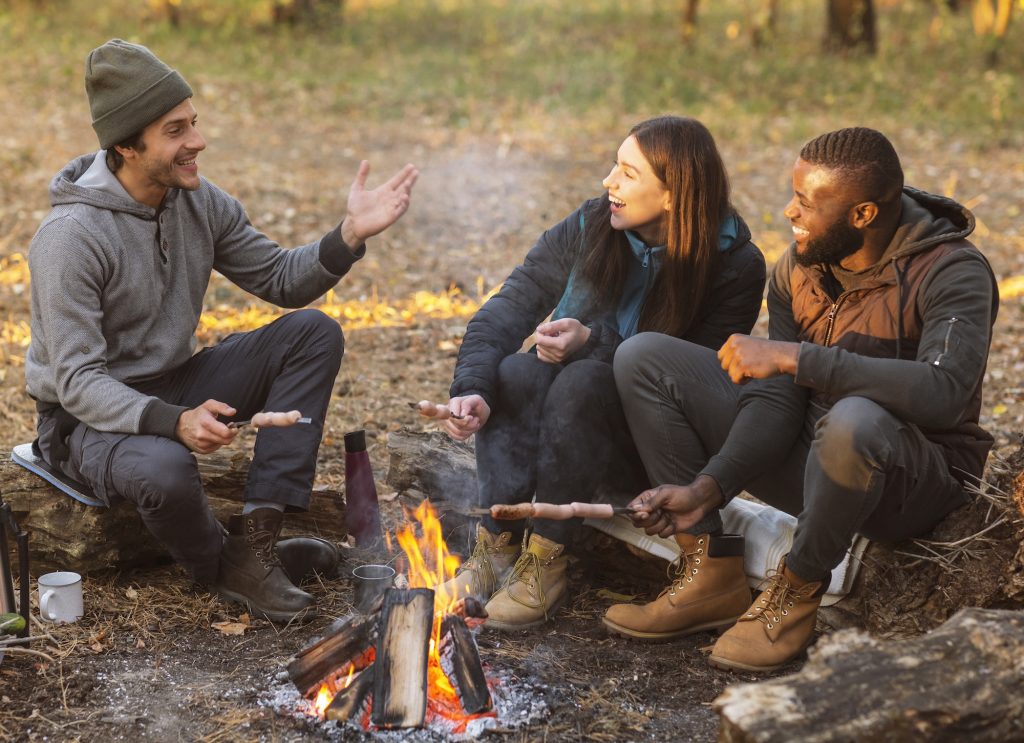 Camping time for mixed race group of friends sitting beside fireplace and frying sausages in forest