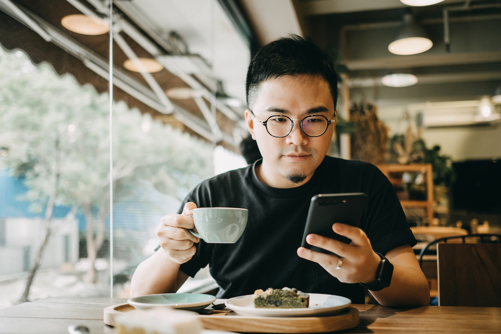 A young man having a relaxed time enjoying coffee and cake with smartphone in cafe