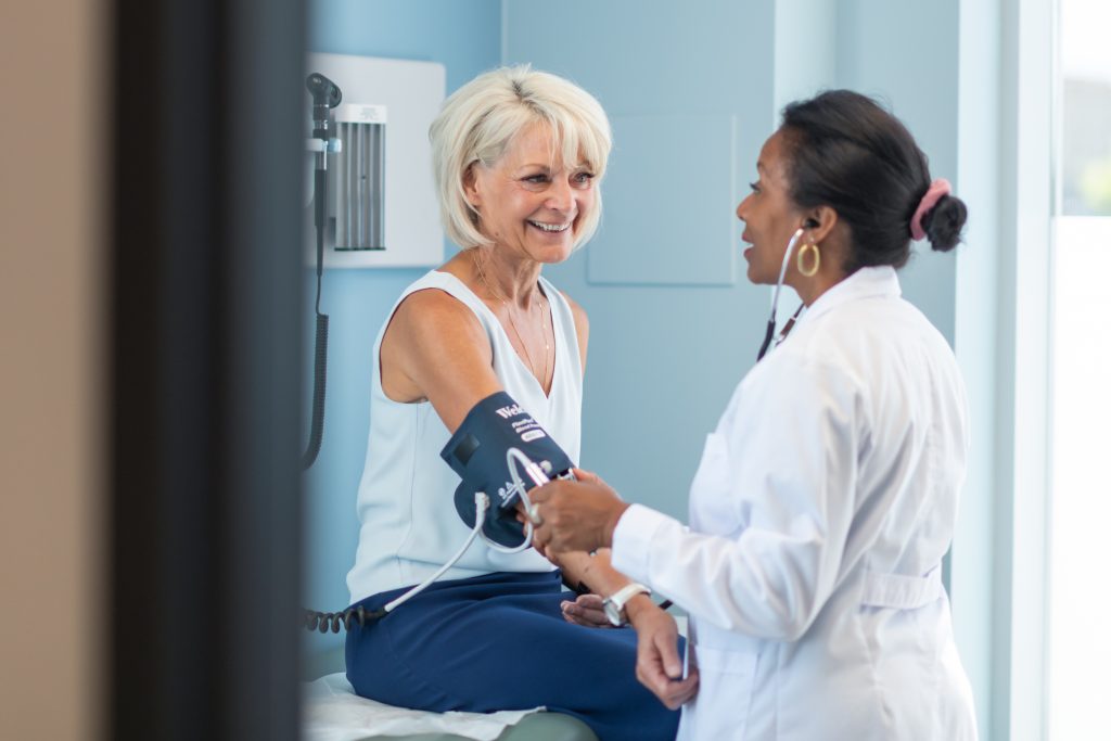 A mature adult woman is at a routine medical appointment. Her healthcare provider is a black woman. The patient is sitting on an examination table in a clinic. She is smiling at the doctor. The doctor is checking the patient's blood pressure.