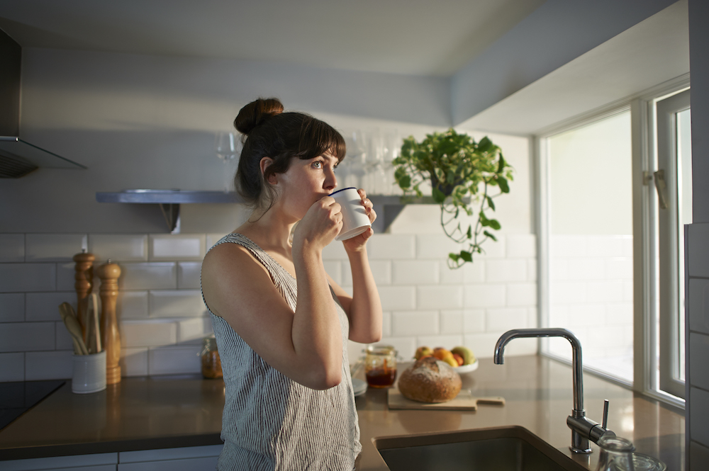 A woman standing in her kitchen drinks from a mug.